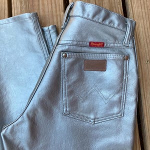 Peut inclure: Un pantalon Wrangler argent&eacute; avec une conception classique &agrave; cinq poches. Le jean pr&eacute;sente un patch en cuir marron et une &eacute;tiquette Wrangler rouge. La finition m&eacute;tallique donne au pantalon un aspect unique et accrocheur.