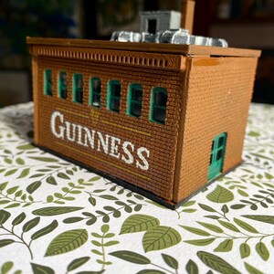 Puede incluir: Un edificio de ladrillo en miniatura con la palabra "Guinness" en letras blancas. El edificio tiene marcos de ventanas verdes y una puerta verde. Se ve una peque&ntilde;a chimenea y detalles del tejado.