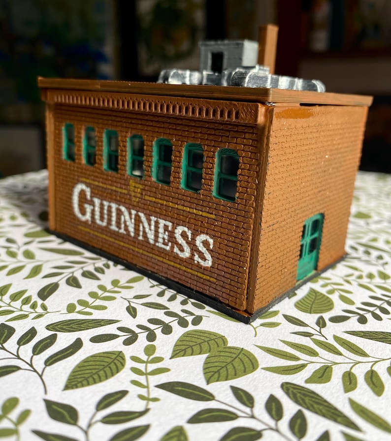 Puede incluir: Un edificio de ladrillo en miniatura con la palabra "Guinness" en letras blancas. El edificio tiene marcos de ventanas verdes y una puerta verde. Se ve una peque&ntilde;a chimenea y detalles del tejado.