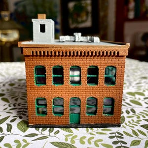 Puede incluir: Un edificio de ladrillo en miniatura con techo plano, chimenea y m&uacute;ltiples ventanas arqueadas. El edificio tiene una fachada de ladrillo marr&oacute;n con marcos de ventanas verdes y una puerta verde. El techo es gris y es un modelo.