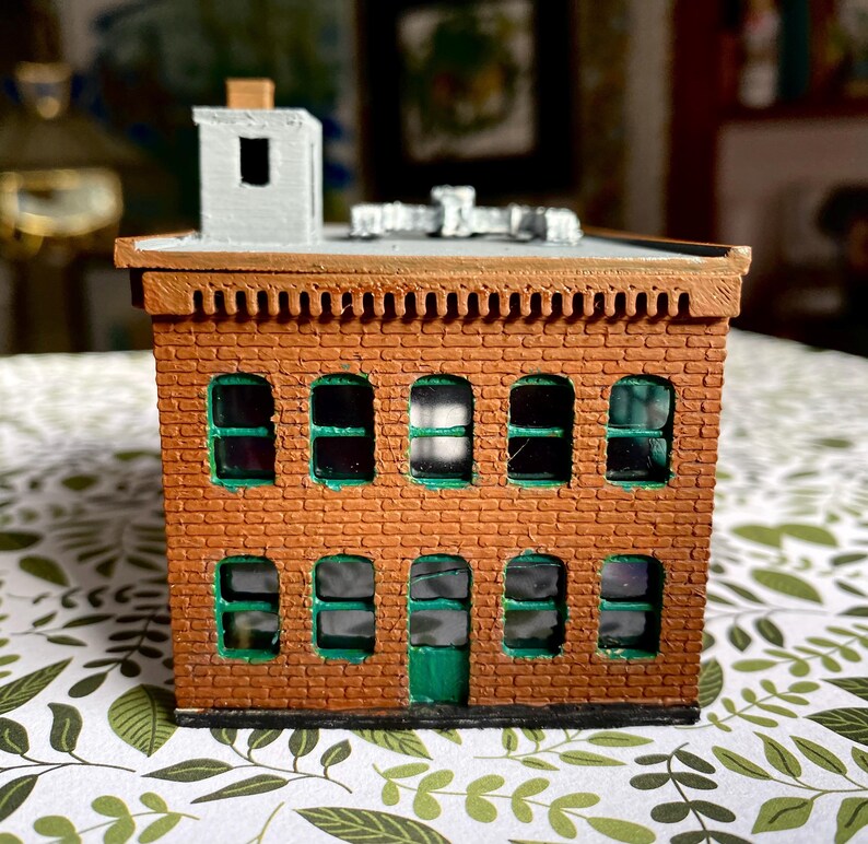 Puede incluir: Un edificio de ladrillo en miniatura con techo plano, chimenea y m&uacute;ltiples ventanas arqueadas. El edificio tiene una fachada de ladrillo marr&oacute;n con marcos de ventanas verdes y una puerta verde. El techo es gris y es un modelo.