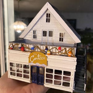 May include: A miniature white building with a gabled roof, featuring a sign that reads "The Canteen." The building has a balcony decorated with colourful buoys and a dark blue door. A black staircase is visible on the side.