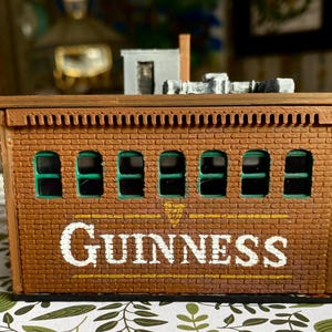 Puede incluir: Un edificio en miniatura de ladrillo marr&oacute;n con la palabra "GUINNESS" en letras blancas. El edificio tiene seis ventanas arqueadas con interiores verdes y una peque&ntilde;a chimenea. El techo tiene un borde decorativo y una peque&ntilde;a estructura en la parte superior.