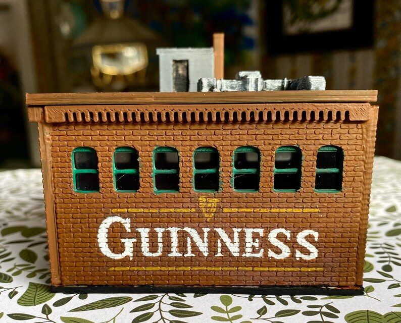 Puede incluir: Un edificio en miniatura de ladrillo marr&oacute;n con la palabra "GUINNESS" en letras blancas. El edificio tiene seis ventanas arqueadas con interiores verdes y una peque&ntilde;a chimenea. El techo tiene un borde decorativo y una peque&ntilde;a estructura en la parte superior.