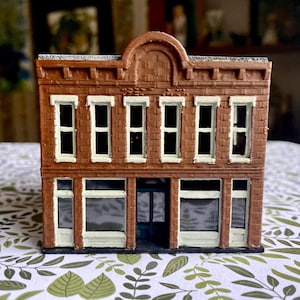 May include: A miniature brown brick building with white window frames and a dark green door. The building has two stories with a decorative arched top. The windows are dark, suggesting a shop or business.