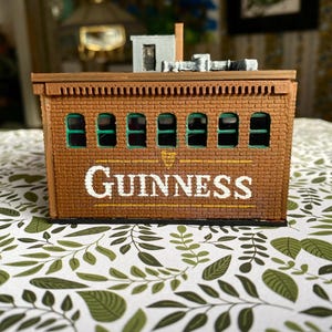 Puede incluir: Un edificio de ladrillo en miniatura con la palabra "GUINNESS" en letras blancas. El edificio tiene ventanas arqueadas con molduras verdes y una peque&ntilde;a estructura en la azotea. La combinaci&oacute;n de colores general es marr&oacute;n y beige.
