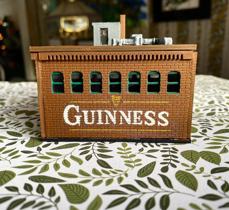 Puede incluir: Un edificio de ladrillo en miniatura con la palabra "GUINNESS" en letras blancas. El edificio tiene ventanas arqueadas con molduras verdes y una peque&ntilde;a estructura en la azotea. La combinaci&oacute;n de colores general es marr&oacute;n y beige.