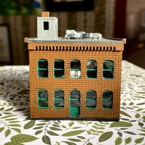 Puede incluir: Un edificio de ladrillo en miniatura con ventanas con marcos verdes y una puerta verde. El edificio tiene un tejado plano con una peque&ntilde;a chimenea y una ventilaci&oacute;n met&aacute;lica. La mamposter&iacute;a es de color marr&oacute;n c&aacute;lido.
