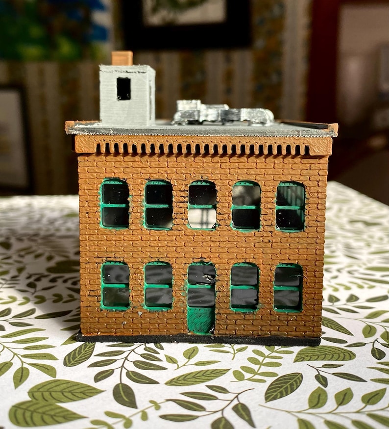 Puede incluir: Un edificio de ladrillo en miniatura con ventanas con marcos verdes y una puerta verde. El edificio tiene un tejado plano con una peque&ntilde;a chimenea y una ventilaci&oacute;n met&aacute;lica. La mamposter&iacute;a es de color marr&oacute;n c&aacute;lido.