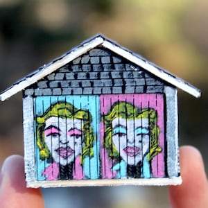 May include: A miniature wooden house with a gray shingled roof and white trim. The front features two painted portraits of a woman with blonde hair, in blue and pink panels. The artwork is held by a person.