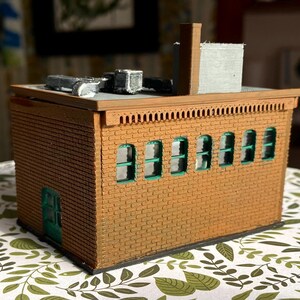 Puede incluir: Un modelo en miniatura de un edificio de ladrillo con un exterior marr&oacute;n, ventanas con marcos verdes y una puerta verde. El tejado tiene una chimenea y unidades de aire acondicionado. El modelo est&aacute; sobre una superficie estampada.