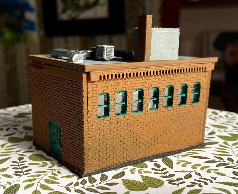 Puede incluir: Un modelo en miniatura de un edificio de ladrillo con un exterior marr&oacute;n, ventanas con marcos verdes y una puerta verde. El tejado tiene una chimenea y unidades de aire acondicionado. El modelo est&aacute; sobre una superficie estampada.