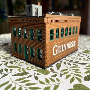 Puede incluir: Un edificio de ladrillo marr&oacute;n en miniatura con marcos de ventanas verdes y la palabra "Guinness" en letras blancas y amarillas. El edificio tiene una peque&ntilde;a estructura gris en el techo. El edificio est&aacute; sobre una superficie estampada.