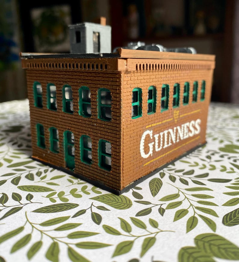Puede incluir: Un edificio de ladrillo marr&oacute;n en miniatura con marcos de ventanas verdes y la palabra "Guinness" en letras blancas y amarillas. El edificio tiene una peque&ntilde;a estructura gris en el techo. El edificio est&aacute; sobre una superficie estampada.