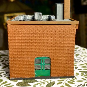 Puede incluir: Modelo en miniatura de un edificio de ladrillo con una puerta verde. El edificio tiene un tejado plano con unidades de ventilaci&oacute;n plateadas y una chimenea marr&oacute;n. La mamposter&iacute;a es de color marr&oacute;n rojizo.