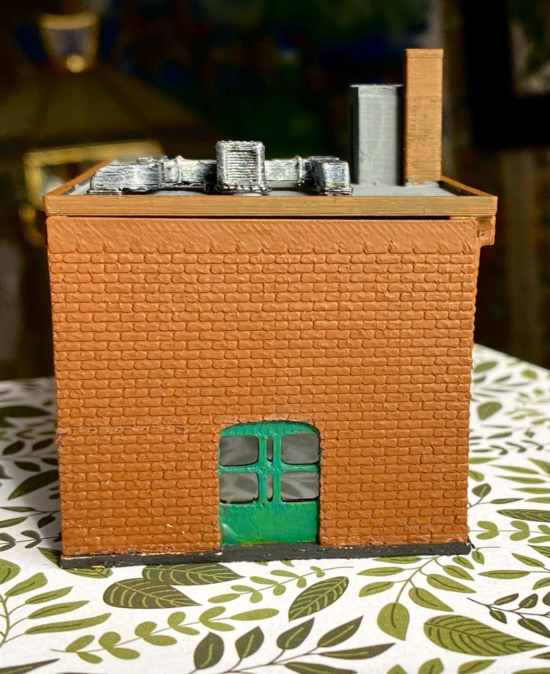 Puede incluir: Modelo en miniatura de un edificio de ladrillo con una puerta verde. El edificio tiene un tejado plano con unidades de ventilaci&oacute;n plateadas y una chimenea marr&oacute;n. La mamposter&iacute;a es de color marr&oacute;n rojizo.