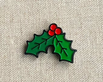 Può includere: Una foglia di agrifoglio verde con tre bacche rosse. La foglia di agrifoglio è bordata di nero.