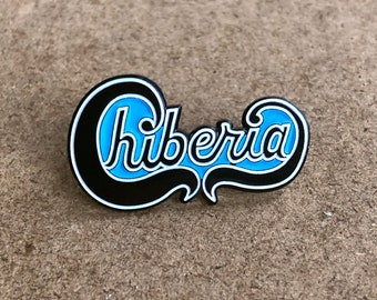 May include: A pin with the word "Chiberia" in a stylized script. The pin features a black border, a light blue background, and white lettering. The pin is likely a collectible or accessory.