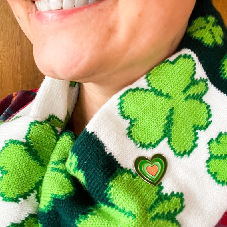 May include: A white and green knitted scarf with a shamrock pattern. A gold heart-shaped pin with a multi-colored heart design is pinned to the scarf.