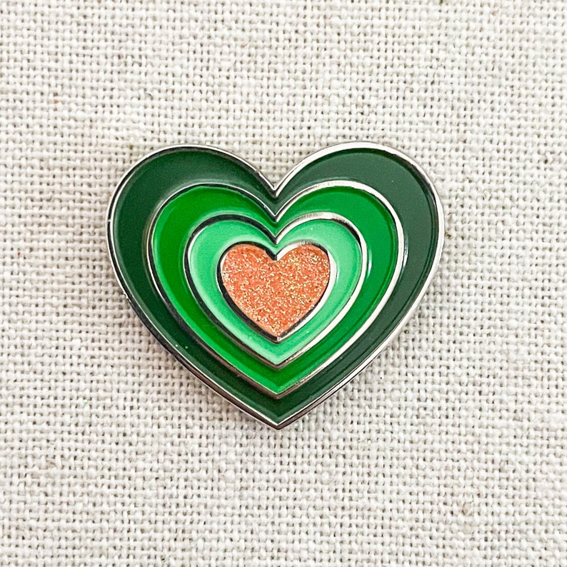 May include: A green heart-shaped enamel pin with three concentric circles and a smaller peach-colored heart in the center.