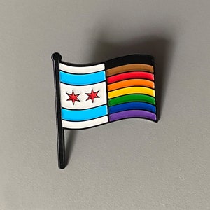 May include: A flag-shaped pin with a black pole and a white border. The flag design features the Chicago city flag on the left and a rainbow-colored LGBTQ+ pride flag on the right. The Chicago flag has two red stars.