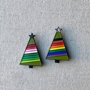 May include: Two Christmas tree-shaped pins. Each pin has a black outline, a green base, and a star at the top. One tree features pink, green, and red stripes, while the other has rainbow stripes.