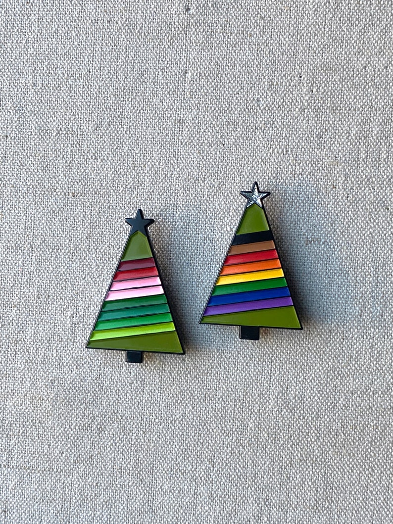 May include: Two Christmas tree-shaped pins. Each pin has a black outline, a green base, and a star at the top. One tree features pink, green, and red stripes, while the other has rainbow stripes.