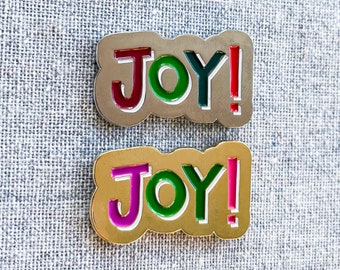 May include: Two enamel pins with the word "JOY!" in a playful font. One pin has a silver-colored border with red, green, and red-colored text. The other pin has a gold-colored border with purple, green, and pink-colored text.