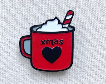 May include: A red enamel pin in the shape of a mug of hot chocolate. The mug has a black heart and the word "xmas" on it. The mug is topped with whipped cream and a red and white striped candy cane.