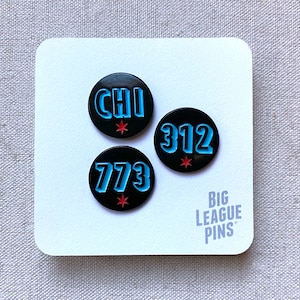 May include: Three black enamel pins with blue and red lettering. The pins feature the words "CHI", "312", and "773". The pins are on a white background with the text "BIG LEAGUE PINS".