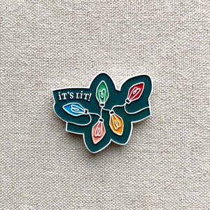 May include: Enamel pin featuring a string of colorful Christmas lights with the text "IT'S LIT!" in white. The lights are in shades of blue, green, red, and orange, set against a dark green background with a silver outline.