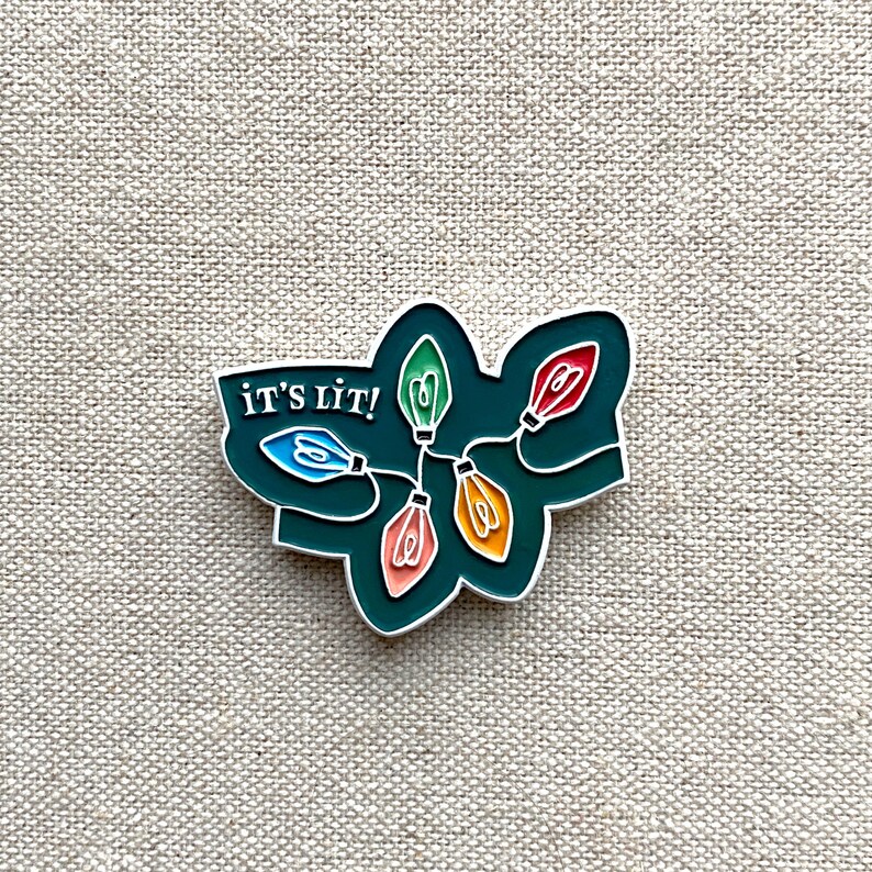 May include: Enamel pin featuring a string of colorful Christmas lights with the text "IT'S LIT!" in white. The lights are in shades of blue, green, red, and orange, set against a dark green background with a silver outline.