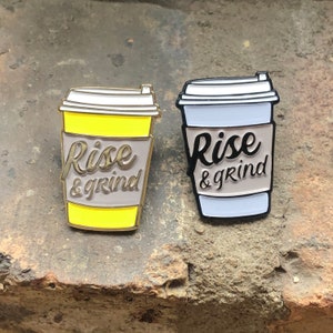 Può includere: Due spille smaltate a forma di tazze da caffè. Entrambe le tazze hanno una fascia beige con il testo "Rise & Grind" in lettere dorate. Una tazza è gialla e l'altra è bianca.