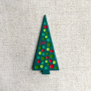 May include: A wooden Christmas tree ornament in a triangular shape. The tree is painted dark green with red and green polka dots. The word "JOY" is written in red near the top of the tree.