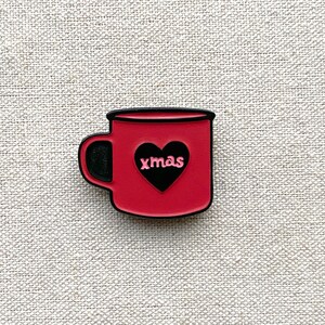 May include: A red mug-shaped charm with a black handle and rim. A black heart shape is in the center of the mug, with the word "xmas" in pink letters inside the heart. The charm is on a textured, light beige surface.