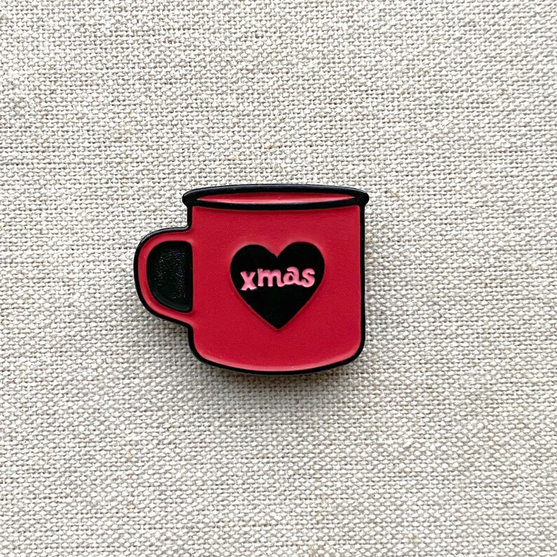 May include: A red mug-shaped charm with a black handle and rim. A black heart shape is in the center of the mug, with the word "xmas" in pink letters inside the heart. The charm is on a textured, light beige surface.