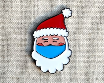 May include: A Santa Claus face pin with a red hat, white beard, and blue face mask. The pin has a black outline and is set against a neutral fabric background. A festive holiday accessory.