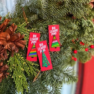 May include: Three red rectangular cards with the text "BIG LEAGUE PINS" and Christmas tree designs. The trees feature rainbow, green, and white colors. The cards are displayed on a wreath of green pine needles, pine cones, and red berries.