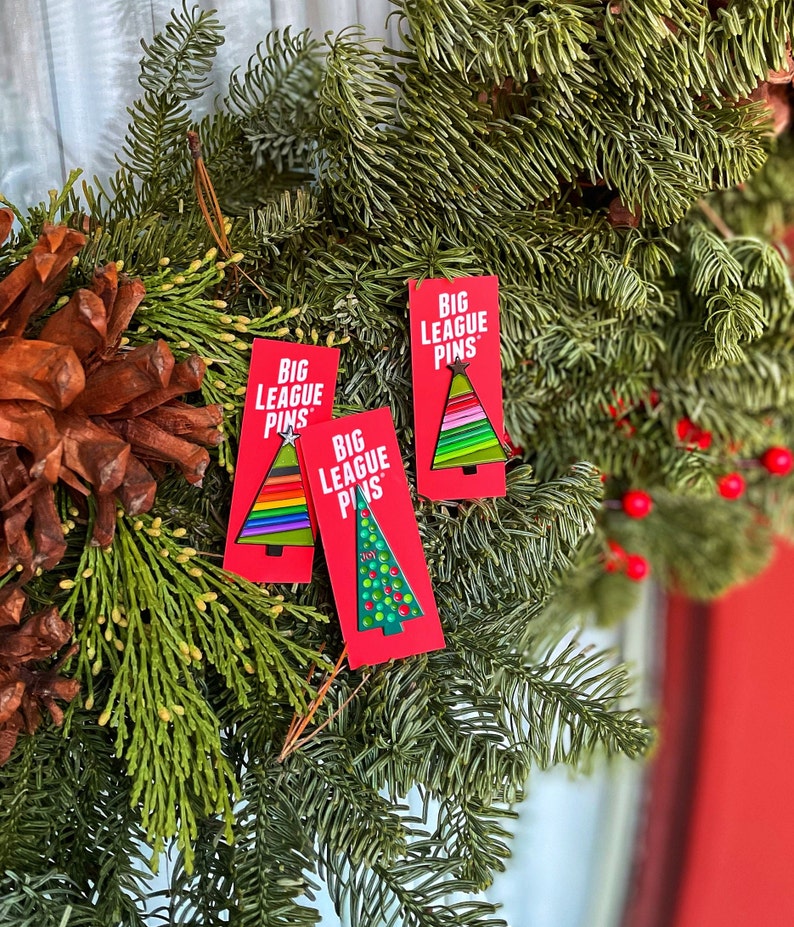 May include: Three red rectangular cards with the text "BIG LEAGUE PINS" and Christmas tree designs. The trees feature rainbow, green, and white colors. The cards are displayed on a wreath of green pine needles, pine cones, and red berries.