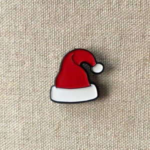 Puede incluir: Pin de esmalte rojo y blanco con forma de gorro de Papá Noel.