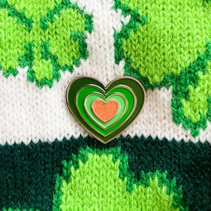 May include: A green, white, and orange heart-shaped pin with a layered design. The pin is on a green and white knitted fabric.