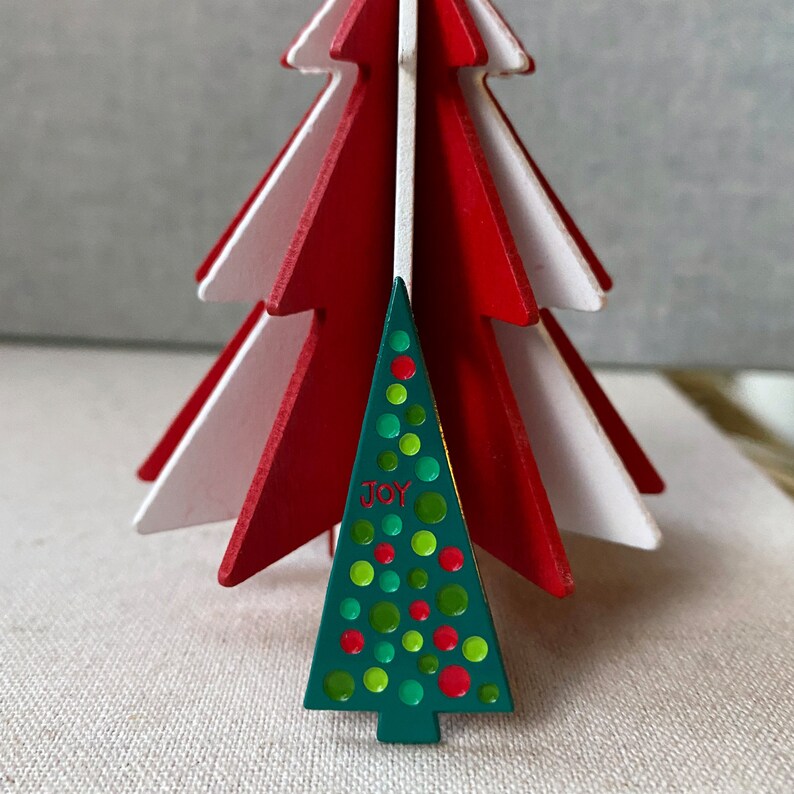 May include: A decorative Christmas tree with a layered design. The tree features red and white wooden layers, with a smaller green tree in front. The green tree is decorated with colorful dots and the word "JOY".