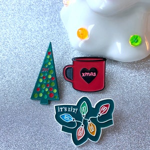 May include: A collection of Christmas-themed enamel pins. Includes a green Christmas tree pin with the word "JOY", a red mug pin with a black heart and the word "xmas", and a green pin with string lights and the phrase "IT'S LIT!"