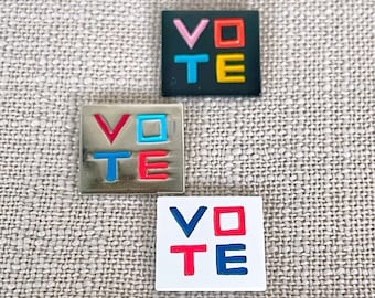 May include: Three square pins with the word "VOTE" spelled out in colorful letters. The pins are black, silver, and white.