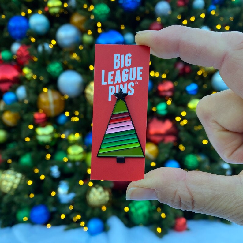 May include: A red rectangular card with the text "BIG LEAGUE PINS" and a Christmas tree pin. The tree pin has a black outline and features green, pink, and red stripes. The background is a blurred Christmas tree with colorful ornaments.