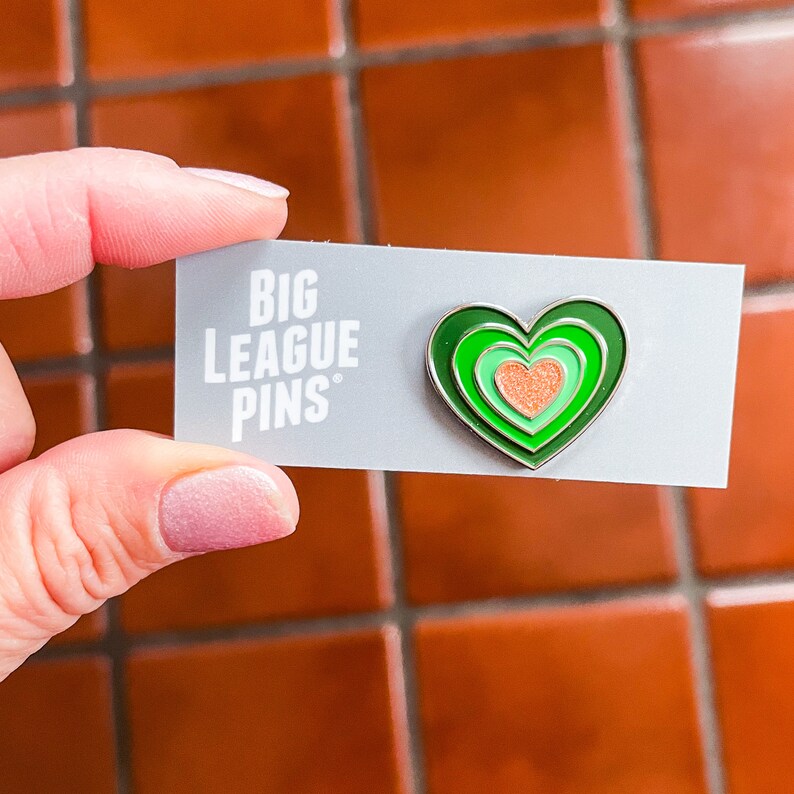 May include: Green and orange enamel pin in the shape of a heart with the text "BIG LEAGUE PINS" on a white card.