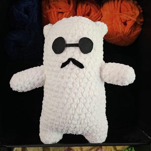 May include: White crocheted plush toy with black sunglasses and a mustache. The toy is sitting in a box with colorful yarn balls.