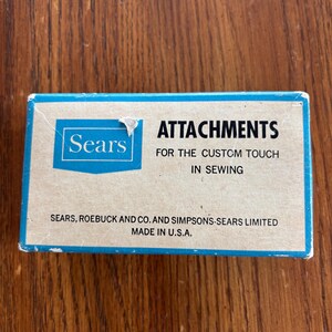 May include: A rectangular cardboard box with a blue border and tan background. The text reads: Sears ATTACHMENTS FOR THE CUSTOM TOUCH IN SEWING. SEARS, ROEBUCK AND CO. AND SIMPSONS-SEARS LIMITED MADE IN U.S.A. The box shows minor wear on the edges.