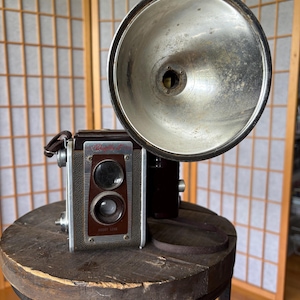May include: Vintage Rolleiflex camera with a large, silver flash attachment. The camera is brown and silver, with a leather strap. It sits on a rustic wooden stool. The camera has the text "Rolleiflex" and "YOKET LENS" on the front.
