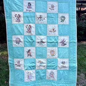 Pode incluir: Colcha de bebê vintage com fundo azul claro e quadrados brancos. Cada quadrado apresenta uma imagem bordada de uma criança ou flor. A colcha está pendurada do lado de fora em um varal.